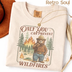 Puede incluir: Camiseta color crema con un gráfico de Smokey Bear señalando, con el texto "ONLY YOU CAN PREVENT WILDFIRES". El diseño incluye una escena de bosque con llamas. También se ven un collar dorado y un sombrero marrón.
