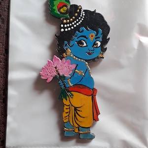 May include: A wooden cutout depicting Krishna in a cartoon style, painted in blue, yellow, and red. The figure holds a bouquet of pink lotus flowers and wears a peacock feather in his hair. The figure is on a white background.