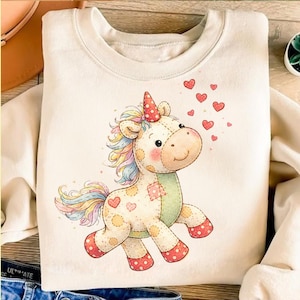 May include: Cream-coloured sweatshirt with a whimsical unicorn design. The unicorn has a colourful mane and tail, a red and white polka-dot horn, and is surrounded by red hearts. The unicorn has a patchwork design with polka dots and hearts.