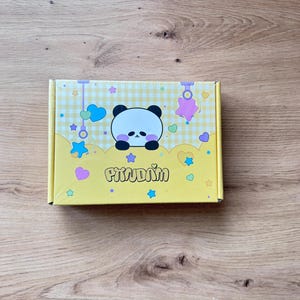 Può includere: Una scatola gialla con un'illustrazione di panda e la parola "Pitudim". La scatola presenta un motivo a quadretti e cuori decorativi, stelle e forme sospese in colori pastello.