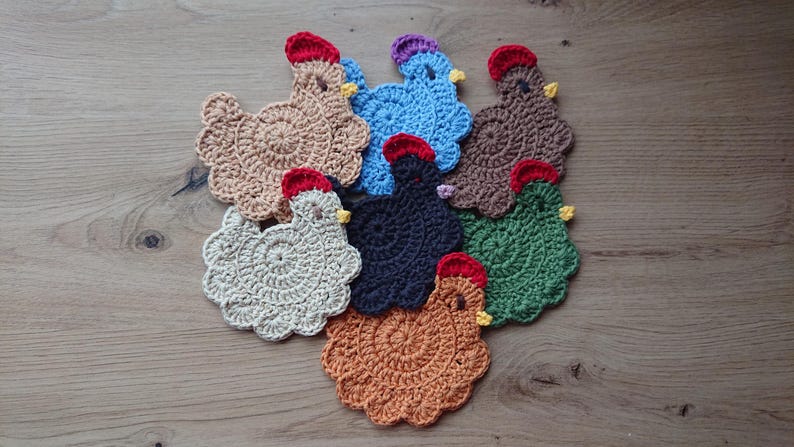 Handmade crochet chiken coasters placed on a wooden desk. The crochet coasters are in different colors- beige, blue, black, brown, light brown, caramel. They have red combs and yellow beaks.