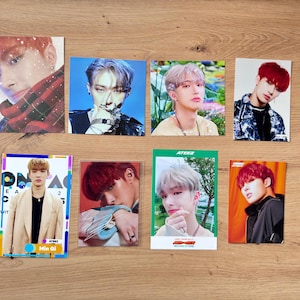 Peut inclure: Huit cartes photo rectangulaires présentant différentes personnes. Les cartes affichent diverses poses et styles, avec une gamme de couleurs de cheveux et de tenues. Une carte porte le nom "Min Gi". Les cartes sont disposées sur une surface en bois.