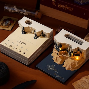 Puede incluir: Dos calendarios de papel decorativos con un diseño de castillo en 3D. Un calendario muestra el mes de junio con los días de la semana. El otro calendario tiene una base azul con la palabra "Hogwarts" impresa. Los calendarios están sobre una superficie de madera.