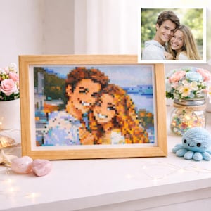 Puede incluir: Un retrato pixelado enmarcado de una pareja sonriente, con un marco de madera. Una foto más pequeña y clara de la pareja está en la esquina superior derecha. El retrato está sobre una superficie blanca, con flores y un pequeño pulpo azul tejido.