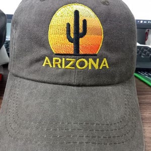 May include: A charcoal grey baseball cap featuring an embroidered design of a black cactus silhouette centred against a circular, gradient sunset background in shades of yellow and orange, with the word ARIZONA stitched in yellow below the graphic.