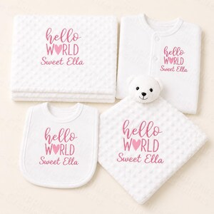 May include: A set of white baby essentials: a blanket, sleepsuit, bib, and comforter. Each piece is embroidered with the pink text "hello WORLD Sweet Ella" and a pink heart. The comforter features a white teddy bear head.
