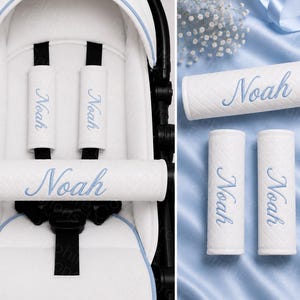 May include: A set of white stroller accessories featuring a quilted texture and the name Noah embroidered in light blue script. The set includes two vertical shoulder strap covers and one horizontal bumper bar cover, all with matching blue trim.