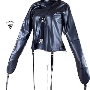 May include: Black faux leather jacket with long sleeves and attached straps. The jacket features a high collar and multiple buckles. The sleeves extend to the hands, with straps at the ends. The Skynza logo is visible in the upper left corner.