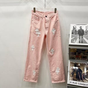 Puede incluir: Vaqueros de mezclilla rosa claro con aplicaciones de cruces blancas. Los vaqueros tienen un diseño clásico de cinco bolsillos y un corte de pierna recta. Las cruces blancas están distribuidas uniformemente en la parte delantera y trasera.