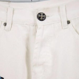 May include: Close-up of white denim pants. The pants feature a black button with a cross design and silver rivets. The design is visible on the front of the pants, showcasing the button and rivet details.