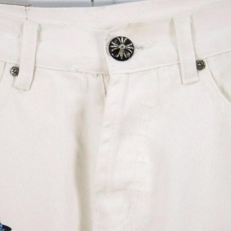 May include: Close-up of white denim pants. The pants feature a black button with a cross design and silver rivets. The design is visible on the front of the pants, showcasing the button and rivet details.