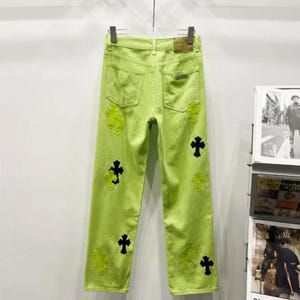 May include: Lime green denim jeans with black and neon green cross appliqu&eacute;s. The jeans have a classic five-pocket design and a straight leg fit. The jeans are hanging on a hanger against a white wall.
