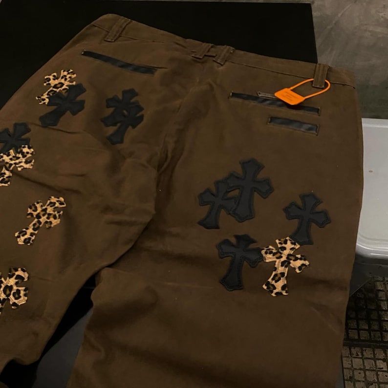 Op de afbeelding: Bruine broek met meerdere kruisapplicaties. Sommige kruisen zijn effen zwart, andere hebben een luipaardprint. De broek heeft zwarte leren accenten en een klein oranje label.