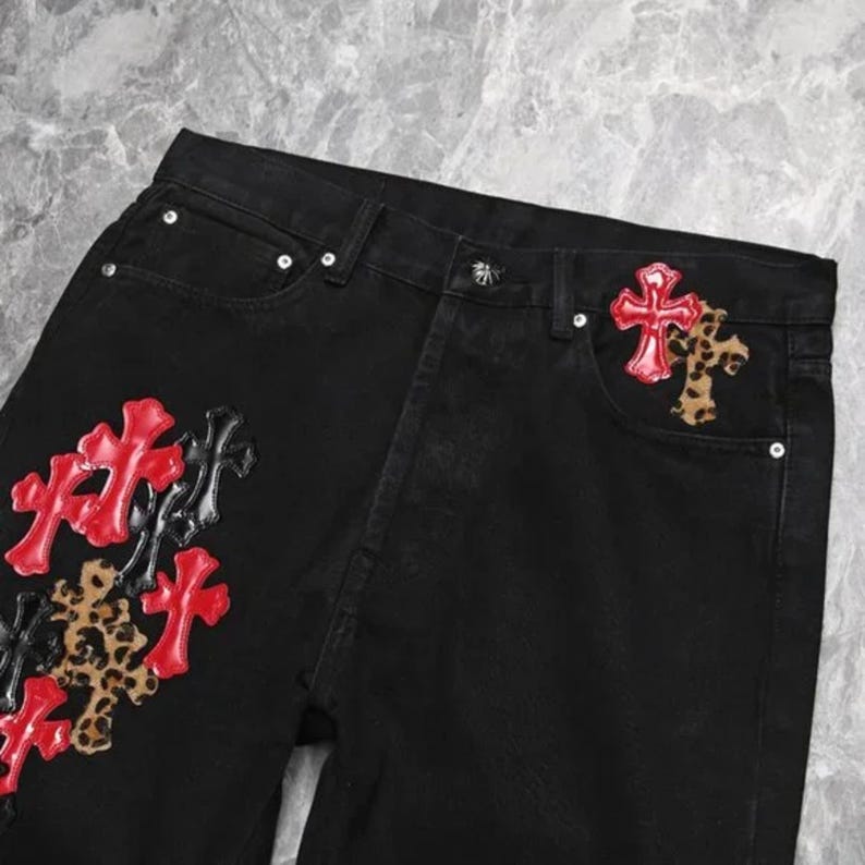 Puede incluir: Vaqueros negros con parches decorativos de cruces. Las cruces son rojas, negras y con estampado de leopardo. Los vaqueros tienen un dise&ntilde;o cl&aacute;sico de cinco bolsillos y cierre de bot&oacute;n y cremallera.