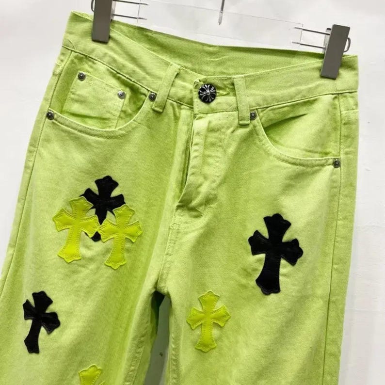 May include: Lime green denim jeans with black and yellow cross appliqu&eacute;s. The jeans have a classic five-pocket design with a button and zipper closure. The crosses are in various sizes and are placed on the front of the jeans.