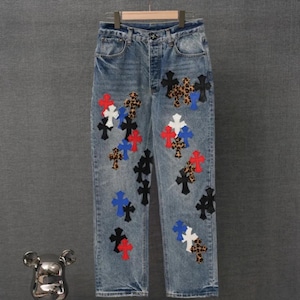 May include: Light blue denim jeans featuring a button fly and a high-waisted fit. The fabric is decorated with numerous cross-shaped patches in various colours including red, white, blue, black, and a brown leopard print pattern scattered throughout.