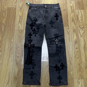 May include: A pair of charcoal gray denim jeans featuring multiple black cross-shaped patches of varying sizes scattered across the front. The jeans include a button fly closure, five pockets, and silver-toned metal rivets at the pocket corners.