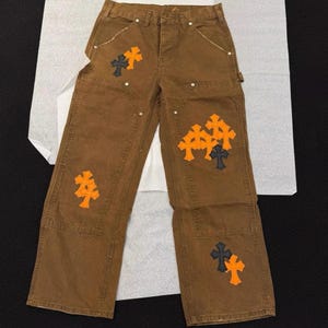 May include: Brown work pants featuring a high-waisted design with a button fly and multiple pockets. The fabric is adorned with several embroidered cross patches in contrasting orange and black colors, distributed across the front legs.