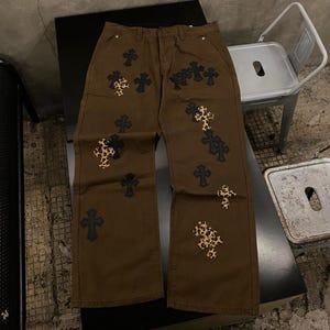 Puede incluir: Pantalones vaqueros marrones con múltiples aplicaciones de cruces. Algunas cruces son negras lisas, mientras que otras presentan un diseño de estampado de leopardo. Los pantalones tienen un diseño clásico de cinco bolsillos y corte recto.