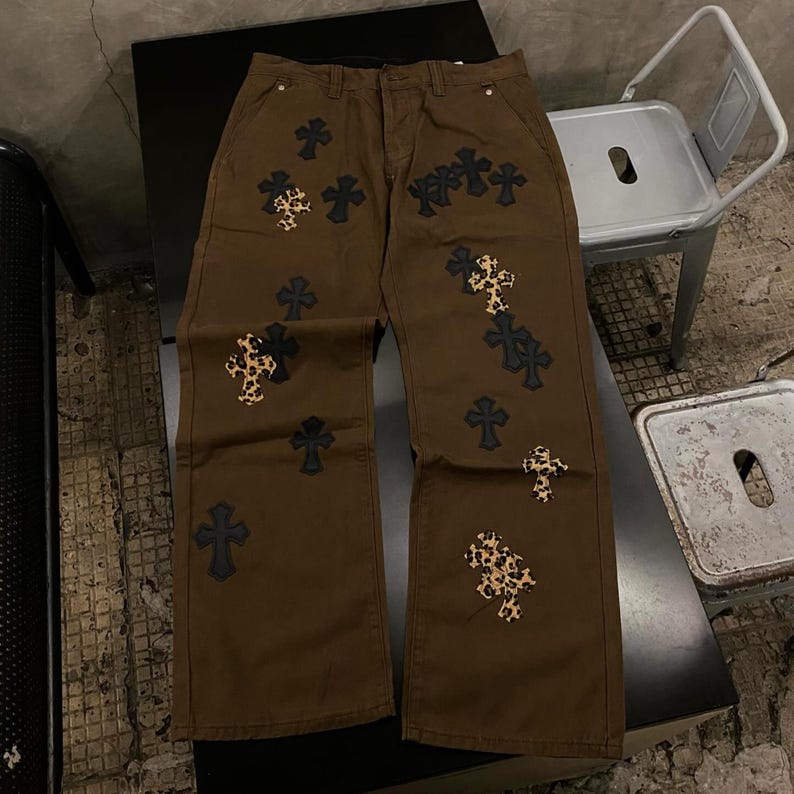 Op de afbeelding: Bruine denim broek met meerdere kruisapplicaties. Sommige kruisen zijn effen zwart, andere hebben een luipaardprint. De broek heeft een klassiek ontwerp met vijf zakken en een rechte pijp.
