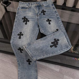 May include: Light blue acid-wash denim jeans featuring a straight-leg cut, a button and zipper fly closure, and multiple black embroidered cross-shaped patches scattered across the front of the legs and the folded pant leg.