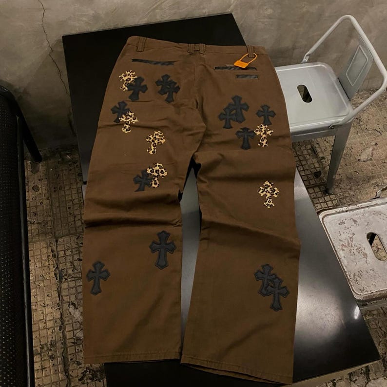 Op de afbeelding: Bruine broek met meerdere kruisontwerpen in zwart en luipaardprint. De broek heeft een relaxte pasvorm en een label aan de achterzak. De kruisen zijn op de voor- en achterkant geborduurd.