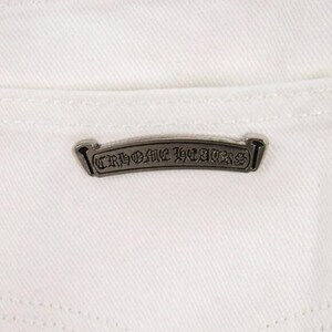May include: Close-up of white denim fabric with a silver metal label. The label is rectangular with the words "CHROME HEARTS" in a gothic font. The label is centered on the back pocket area.
