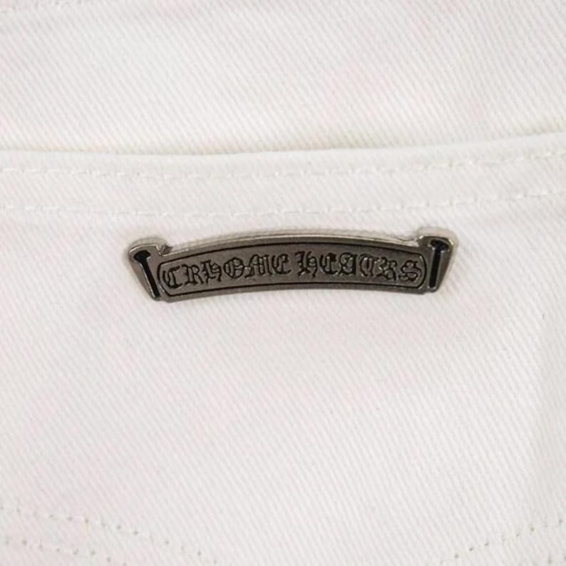 May include: Close-up of white denim fabric with a silver metal label. The label is rectangular with the words "CHROME HEARTS" in a gothic font. The label is centered on the back pocket area.