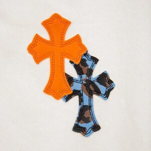 May include: Two decorative crosses on a white background. One cross is orange with a stitched outline, and the other is blue with a brown and black camouflage pattern. The crosses are overlapping.