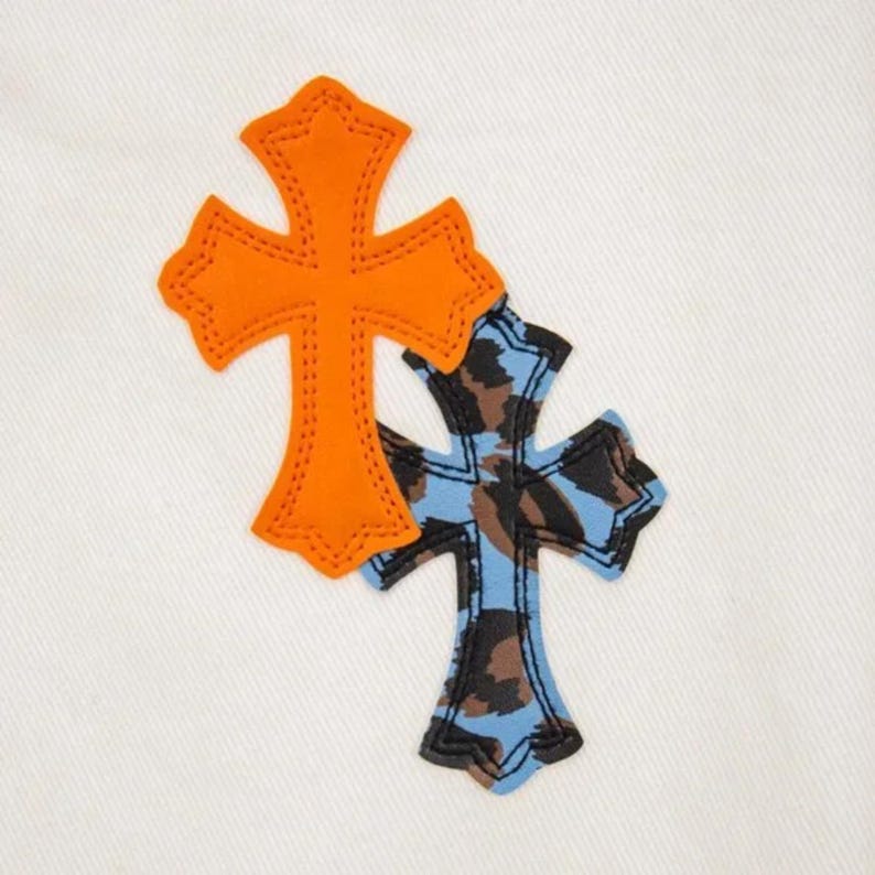 May include: Two decorative crosses on a white background. One cross is orange with a stitched outline, and the other is blue with a brown and black camouflage pattern. The crosses are overlapping.