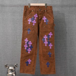 May include: Brown pants featuring a series of purple and blue cross-shaped patches scattered across the front. The pants include a button closure, a zipper fly, and multiple pockets, with visible stitching details along the seams and legs.