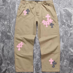Pode incluir: Calças cáqui com vários patches de cruz em rosa e padrões xadrez em preto e branco. As calças têm um design clássico com bolsos e fecho de botão.