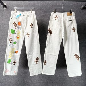 May include: Two pairs of white denim jeans. One pair has colorful graffiti-style designs and cross patterns. The other pair has cross patterns. Both pairs are hanging on a metal rod.