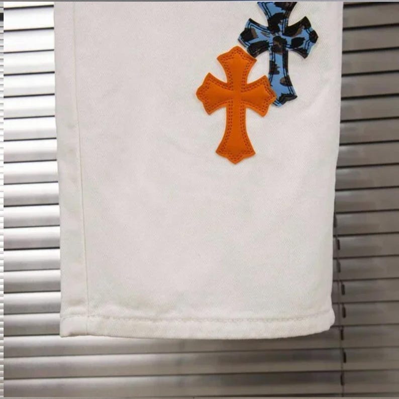 May include: White denim pants with two decorative crosses. One cross is orange, and the other is blue with a leopard print pattern. The crosses are stitched onto the pants.