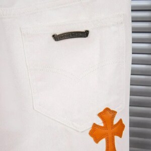 May include: Close-up of white denim jeans featuring a back pocket with a decorative metal emblem and an orange cross-shaped leather patch. The jeans have a classic five-pocket design.