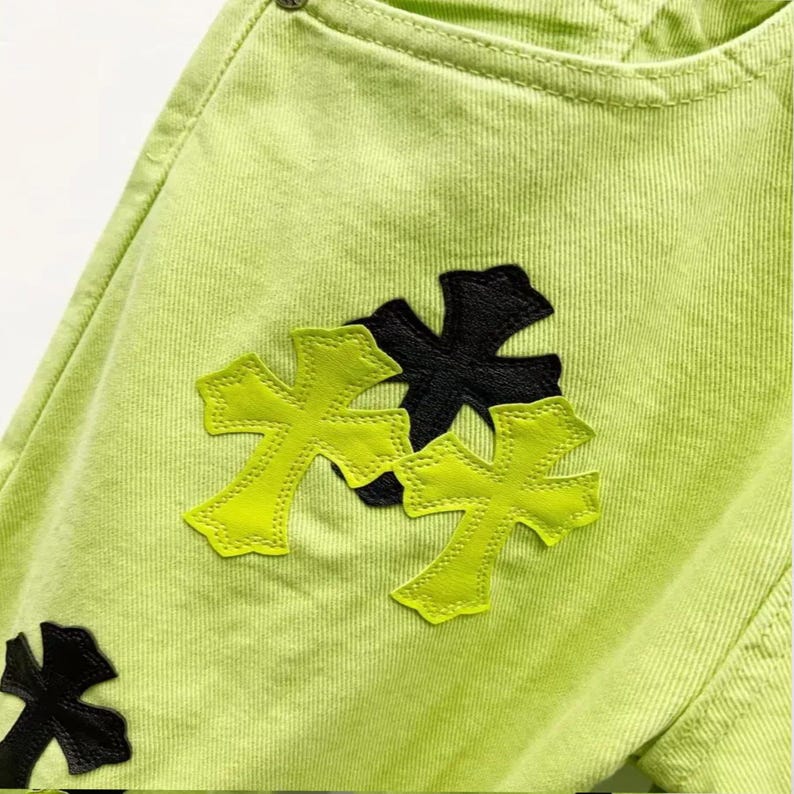 May include: Close-up of lime green denim fabric with black and neon yellow cross-shaped appliqu&eacute;s. The design features a pocket detail and a button. The crosses are stitched with a contrasting yellow thread.