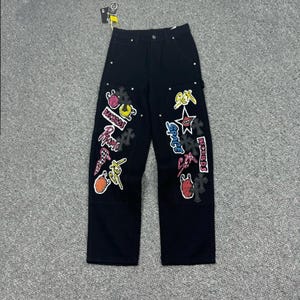 Peut inclure: Pantalon en jean noir à taille haute avec fermeture à bouton et rivets métalliques. Les jambes sont ornées de divers écussons brodés colorés, incluant les textes "SEX", "RECORDS", "PROMO" et "SPACE" ainsi que des motifs d'étoiles et de croix.