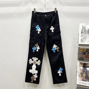 Op de afbeelding: Zwarte denim broek met meerdere kruisapplicaties in wit, blauw en luipaardprint. De broek heeft een klassiek ontwerp met vijf zakken en hangt aan een metalen hanger. De kruisen zijn van verschillende groottes en op de voorkant geplaatst.