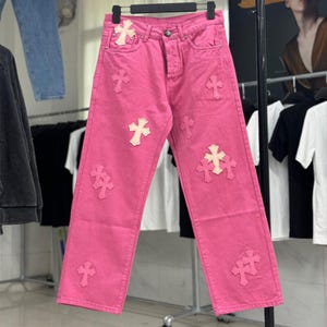 Puede incluir: Vaqueros de mezclilla en color rosa brillante con múltiples parches en forma de cruz de color claro cosidos en la parte delantera. El pantalón incluye una bragueta de botones, cinco bolsillos y remaches metálicos plateados.