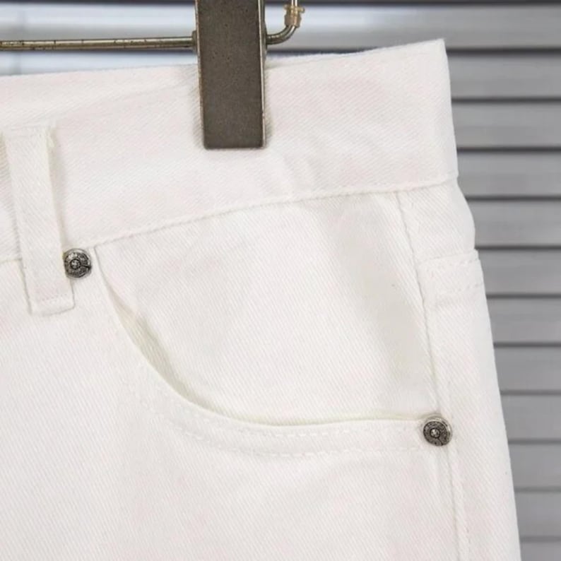 May include: Close-up of white denim jeans. The jeans feature a classic five-pocket design with a button and rivets. The fabric appears to be a sturdy cotton blend. The jeans are hanging on a metal hanger.