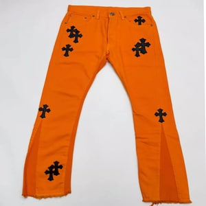 Puede incluir: Vaqueros de mezclilla naranja brillante con aplicaciones de cruces negras. Los vaqueros tienen un diseño de pierna acampanada con una abertura en el dobladillo. Los vaqueros están hechos de un material de mezclilla resistente.