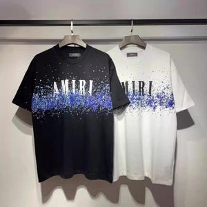 Könnte beinhalten: Zwei kurzärmelige T-Shirts, eines schwarz und eines weiß, mit dem Wort "AMIRI" in weißen Blockbuchstaben. Beide haben ein blau-weißes Spritzmuster auf der Brust. Die T-Shirts hängen an einer Kleiderstange.