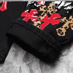 Puede incluir: Primer plano de unos vaqueros negros con cruces rojas y apliques de cruces con estampado de leopardo. Los vaqueros est&aacute;n doblados en el bajo, mostrando el denim de lavado oscuro y los detalles decorativos. El dise&ntilde;o es atrevido y llamativo.