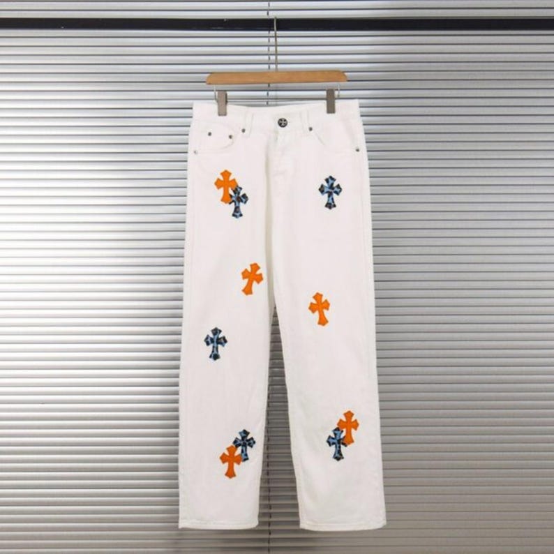 May include: White denim pants with multiple orange and blue cross designs. The pants are hanging on a wooden hanger against a neutral background. The crosses are embroidered on the front of the pants.