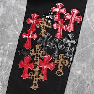 Puede incluir: Pantalones vaqueros negros con m&uacute;ltiples aplicaciones de cruces en rojo, negro y estampado de leopardo. Las cruces est&aacute;n dispuestas en un patr&oacute;n disperso sobre la tela. Las cruces rojas tienen un acabado brillante, mientras que las negras son mate.