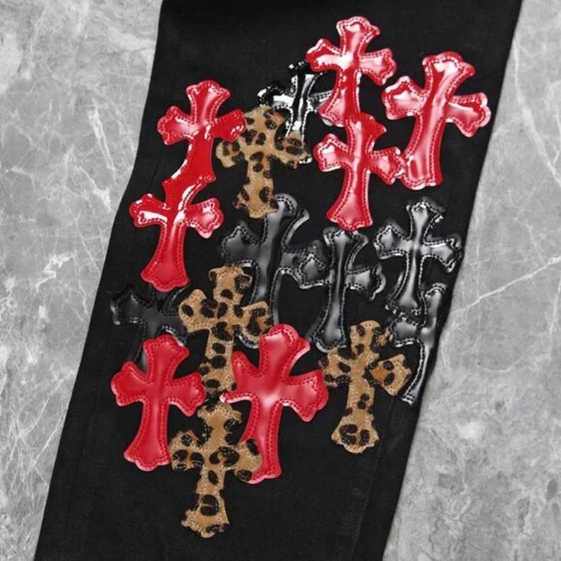 Puede incluir: Pantalones vaqueros negros con m&uacute;ltiples aplicaciones de cruces en rojo, negro y estampado de leopardo. Las cruces est&aacute;n dispuestas en un patr&oacute;n disperso sobre la tela. Las cruces rojas tienen un acabado brillante, mientras que las negras son mate.