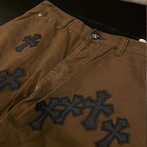 Op de afbeelding: Bruine broek met zwarte kruisapplicaties. De broek heeft een knoopsluiting en riemlussen. De kruizen zijn op de stof gestikt. De broek is gemaakt van een stevig materiaal.