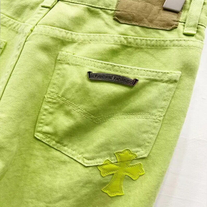 May include: Close-up of lime green denim jeans. The jeans feature a back pocket with a Chrome Hearts logo and a yellow cross patch. A brown leather patch is visible near the waistband.