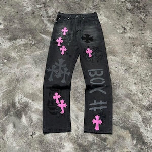 Può includere: Jeans in denim nero a gamba dritta con chiusura a bottoni, decorati con molteplici toppe a forma di croce in rosa acceso. Il tessuto presenta un lavaggio sbiadito, con ulteriori motivi a croce grigio scuro e la scritta "BOX" sulla gamba destra.