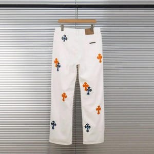 May include: White denim pants with a straight leg cut, adorned with orange and blue cross-shaped embroidery. The pants are hanging on a wooden hanger against a neutral background.